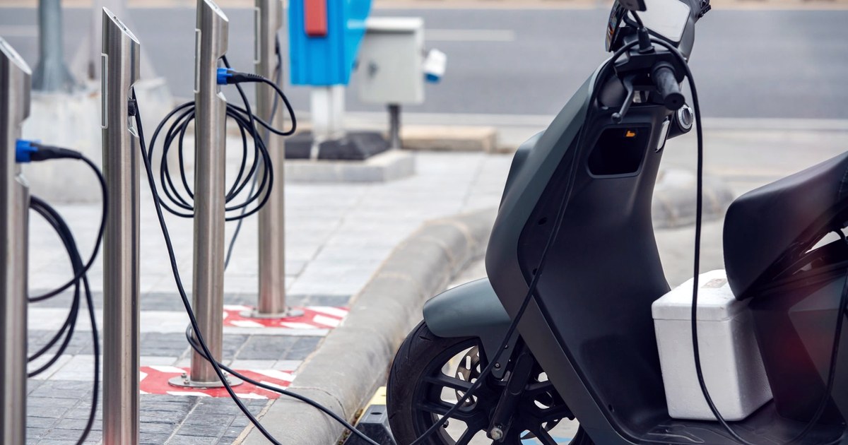A dark electric scooter plugged into a row of steel charging posts on a city pavement