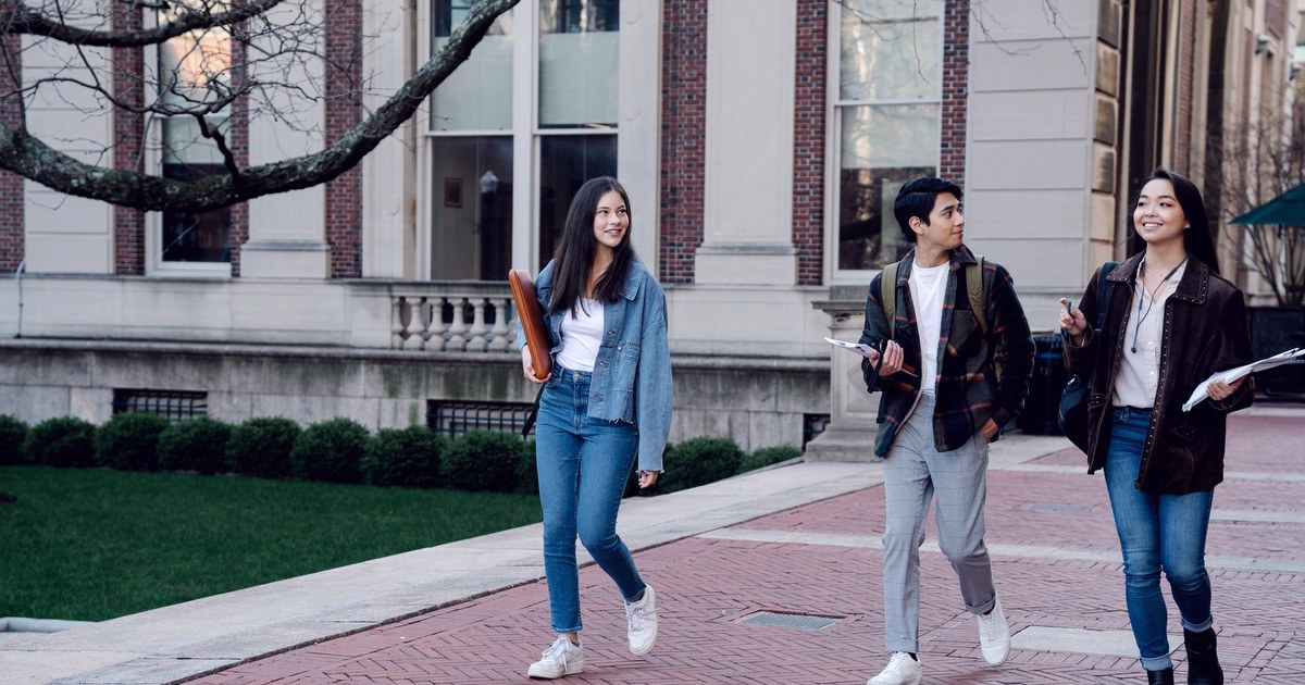 Three international students carrying notebooks and a laptop bag, walking and talking on a university campus courtyard