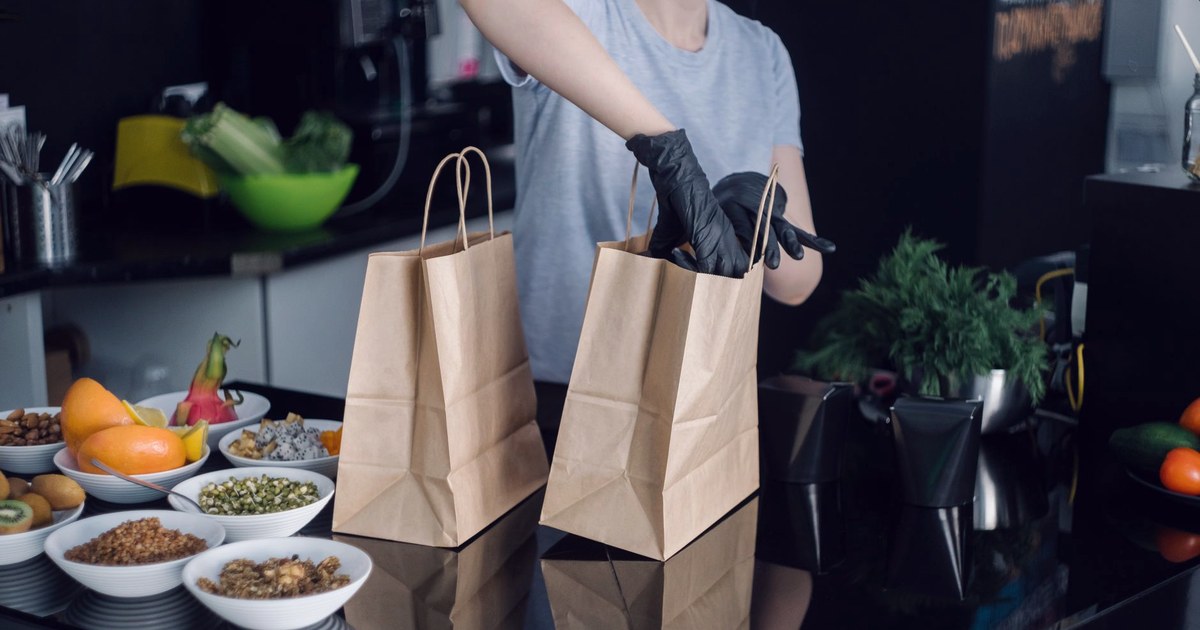 A gloved worker packing fresh produce into paper delivery bags, with bowls of fruit and vegetables on the counter