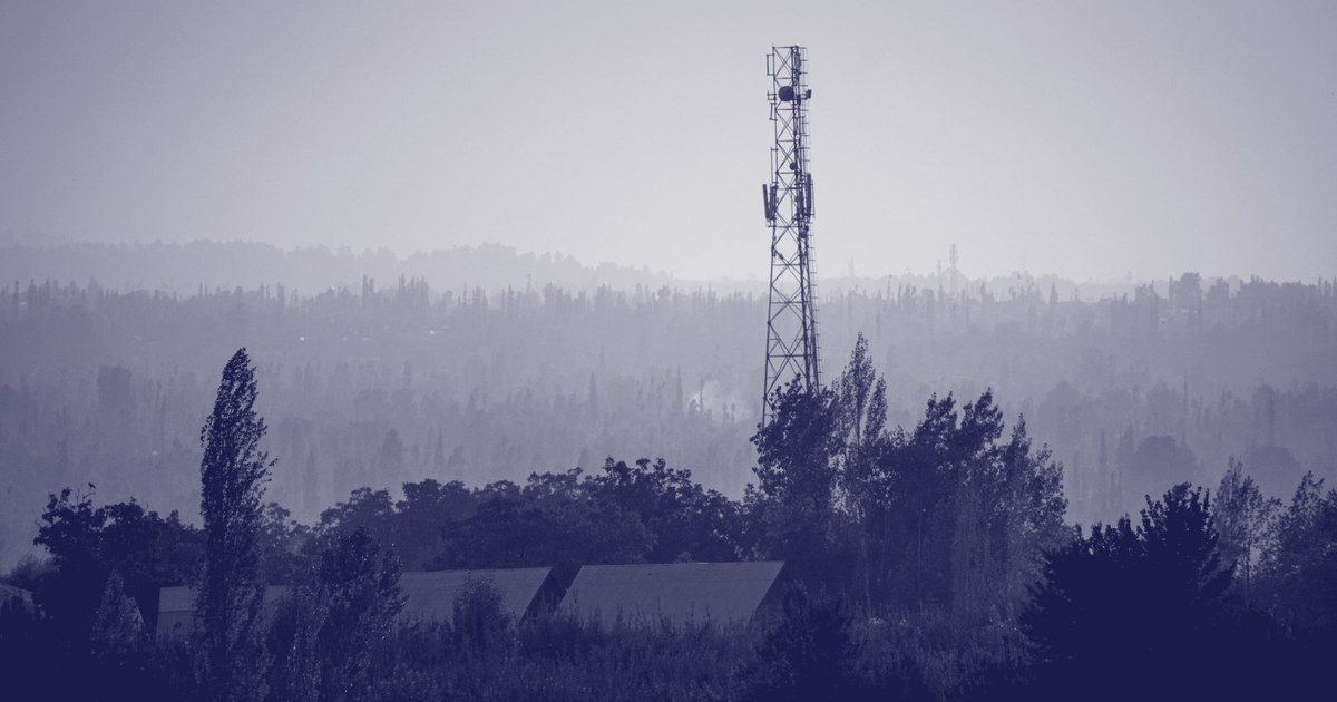 A mobile network tower silhouetted against a hazy sunset above trees and rooftops in a rural Indian landscape