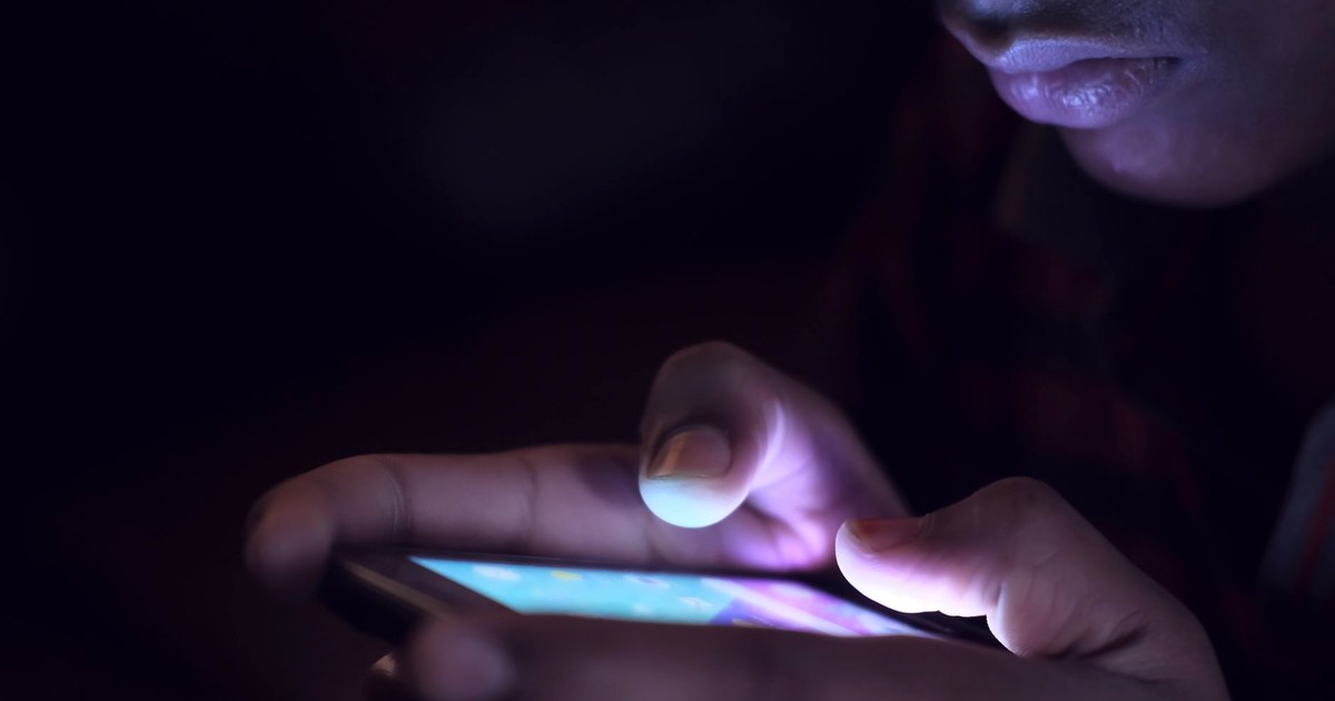 Close-up of a person's hand touching an illuminated smartphone screen in a dark room, only the edge of a face visible in the glow