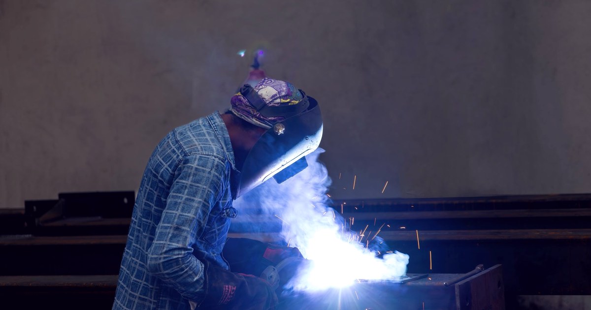 A welder in a plaid shirt and protective mask working on a steel beam in a dim industrial workshop, sparks and blue-white arc light flaring