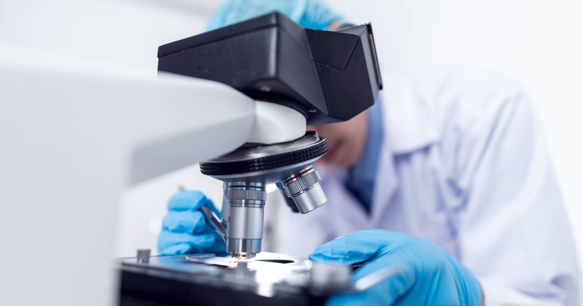 A researcher wearing blue gloves and a face mask examines a sample through a microscope in a laboratory
