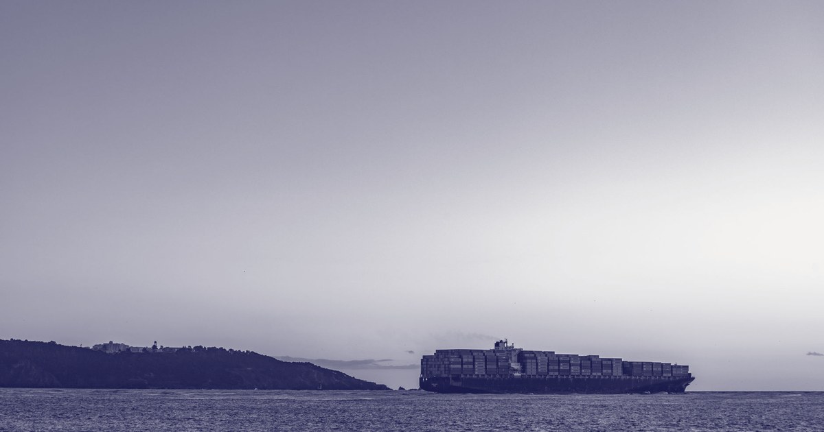 A container ship silhouetted against a dusk sky, sailing past a headland