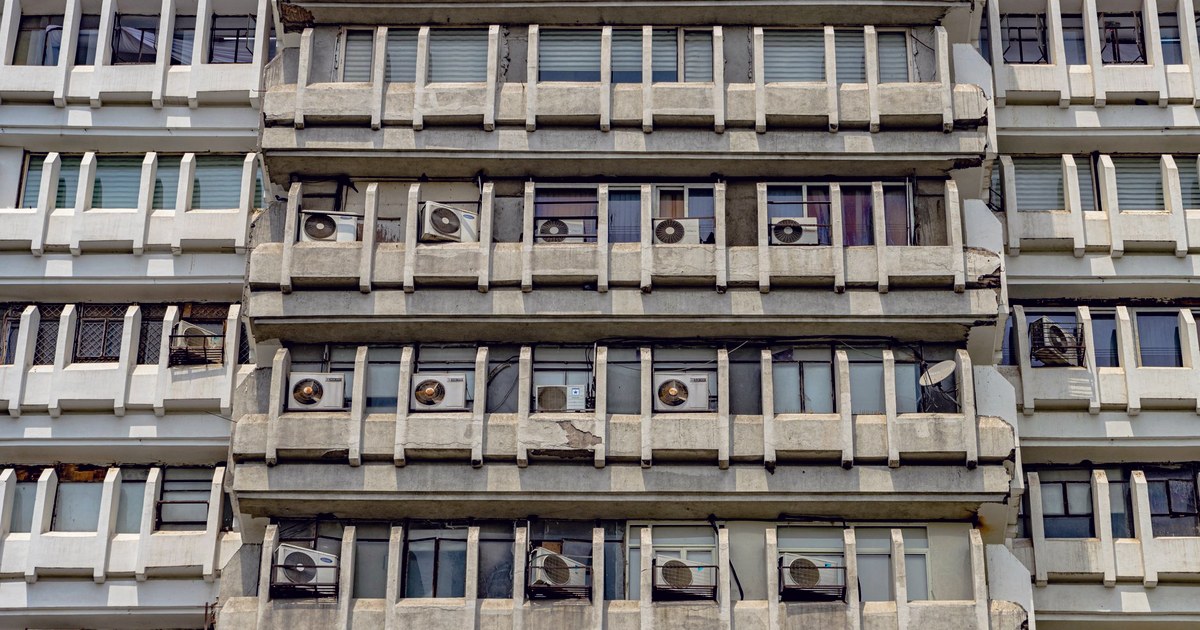 A dense multi-storey residential building facade in New Delhi, with air conditioning units bolted onto balconies and a visible structural crack running down one corner