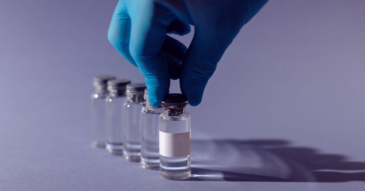 A gloved hand lifting a glass vaccine vial with a blank label from a row of identical vials