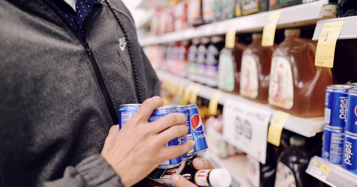 A shopper's hand holding Pepsi cans while selecting bottled beverages from a grocery store shelf