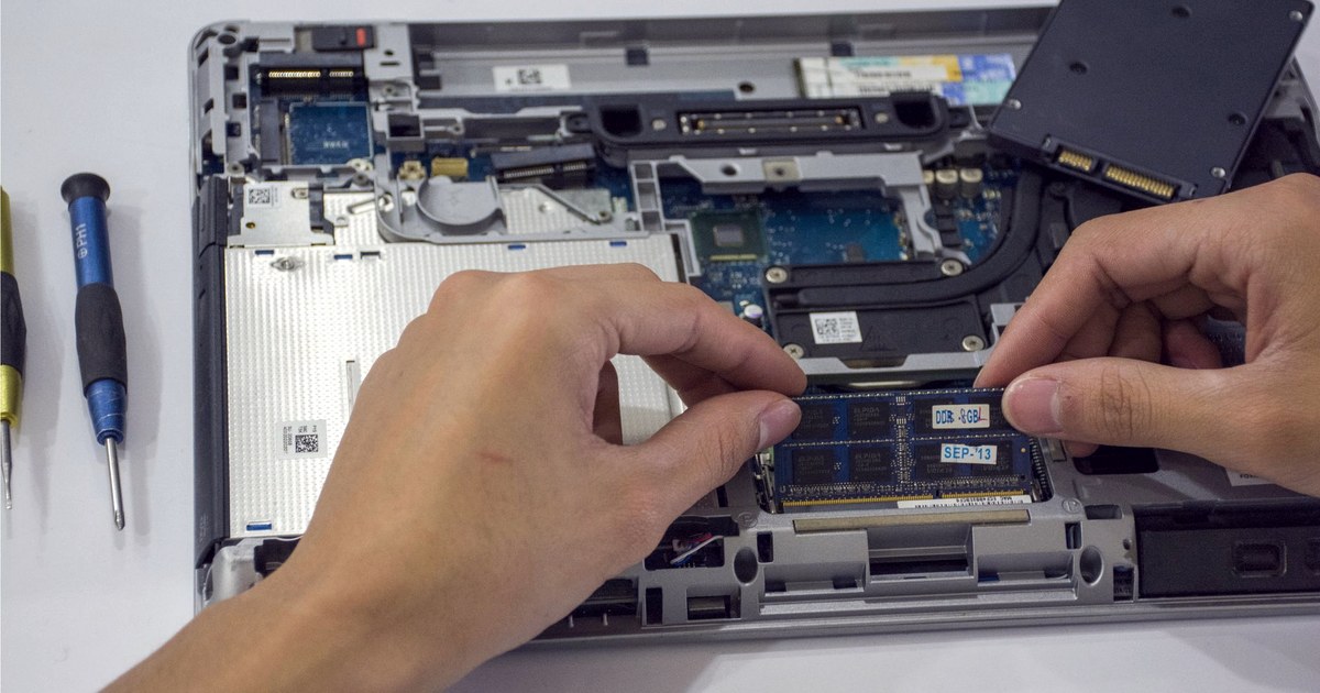 Close-up of hands fitting a memory module into the open base of a laptop, with a screwdriver beside it