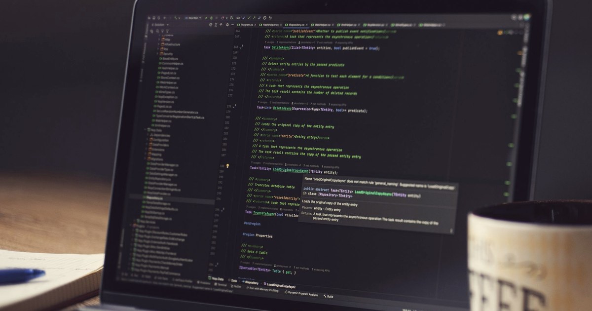 A laptop screen showing lines of source code in a dark code editor, next to a notebook, pen and coffee mug on a wooden desk