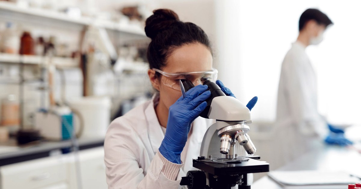 A lab scientist in a white coat and blue gloves examines a sample through a microscope, with a colleague working at a bench in the background