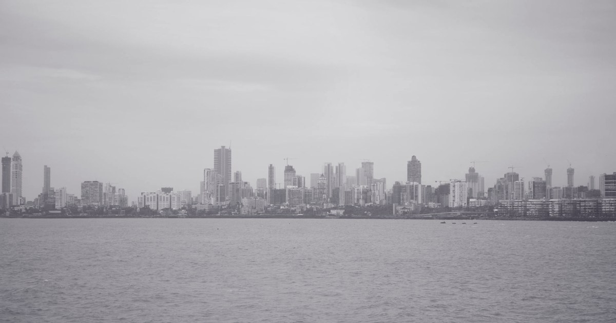 A grayscale view of the Mumbai skyline seen across the harbor, with the city's high-rise towers along the waterfront