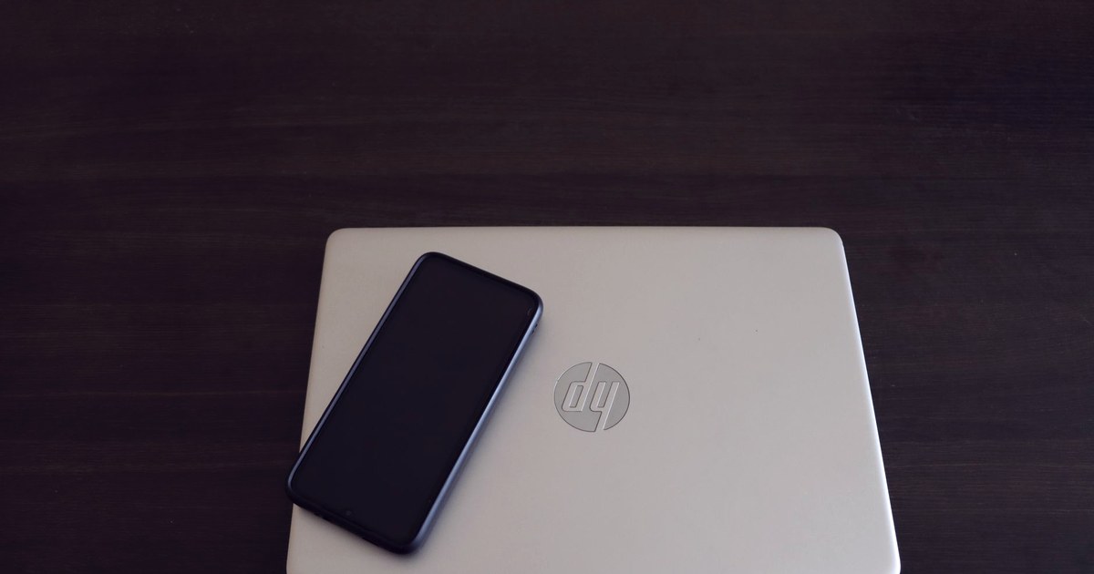 A closed HP laptop with its logo visible, a smartphone resting on top, on a dark wooden desk