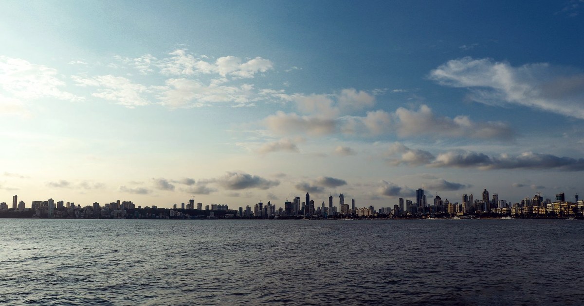 The Mumbai skyline seen across the water from the waterfront, under a partly cloudy sky