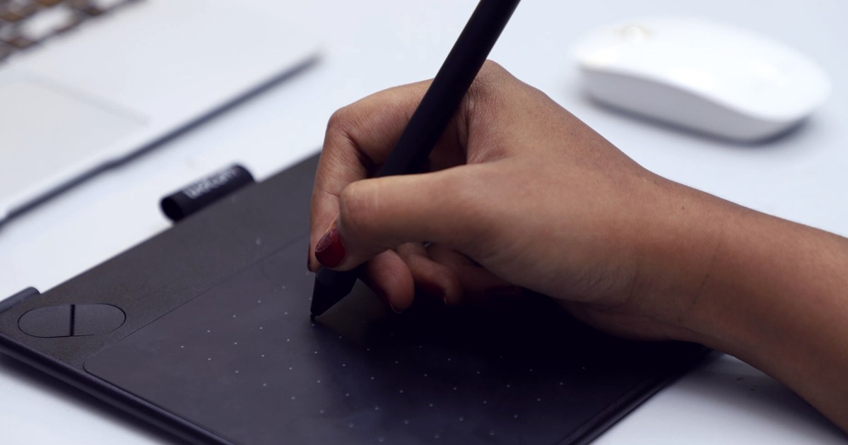 A hand with red nail polish holds a stylus on a graphics tablet next to a laptop, evoking design and creative work