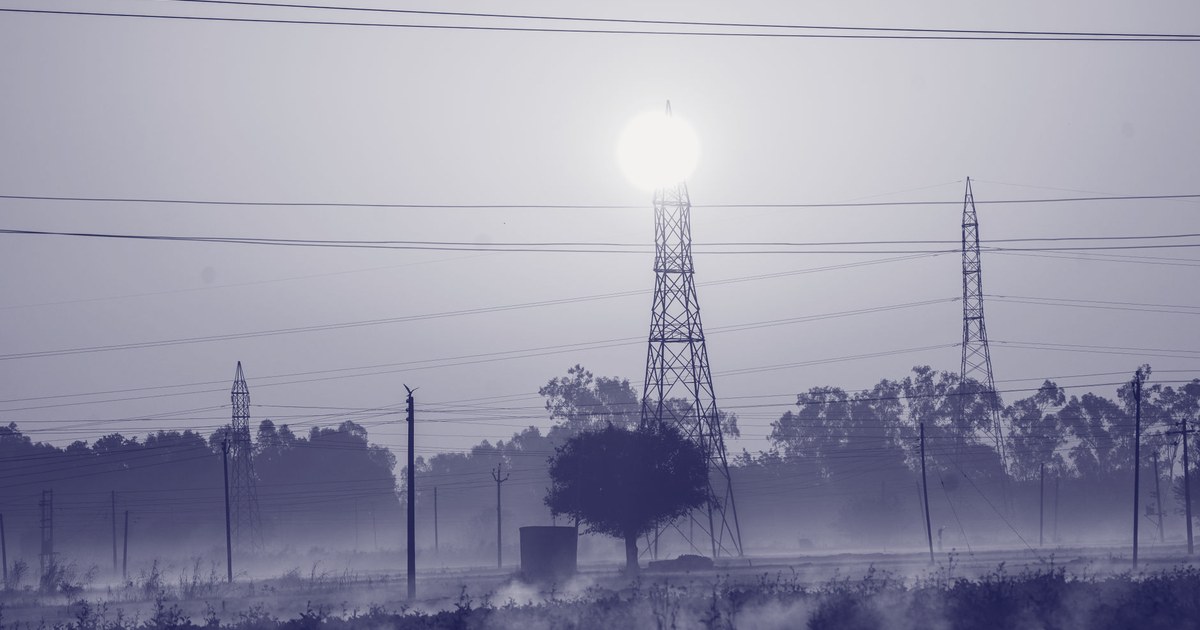 Electricity transmission towers silhouetted against a hazy sunrise over an open field in Punjab, India, power lines converging toward the sun