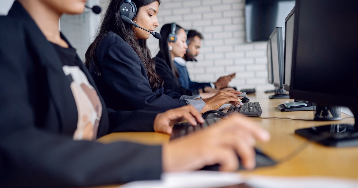 Four office workers wearing telephone headsets at a row of computer desks in a modern call center, one woman in a dark blazer focused on typing