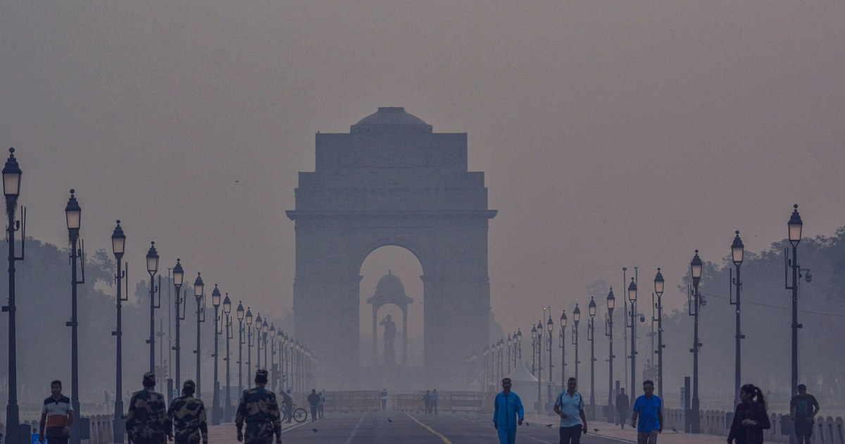 India Gate in New Delhi shrouded in thick smog at dawn, with soldiers and pedestrians walking along the avenue lined with lampposts
