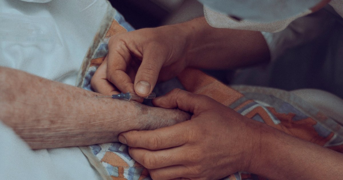 A nurse inserting an intravenous cannula into an elderly patient's forearm, the patient's hand resting in the caregiver's hands