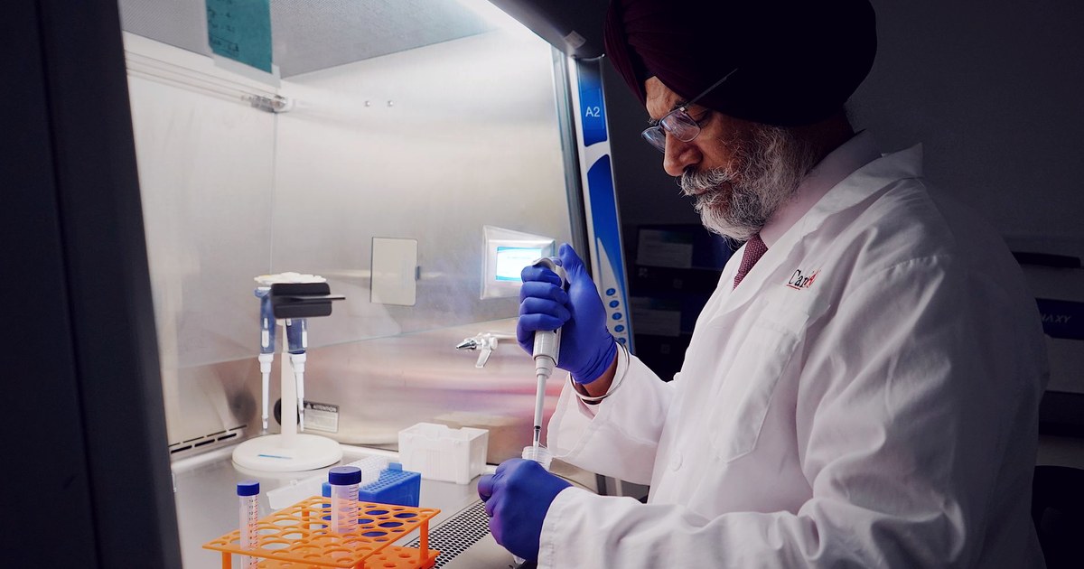 A turbaned Sikh scientist in a white lab coat and blue gloves using a pipette at a laboratory workstation