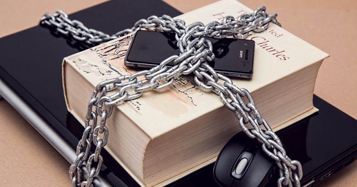 A smartphone, book, and laptop bound together with heavy metal chains, symbolizing restricted access to digital information
