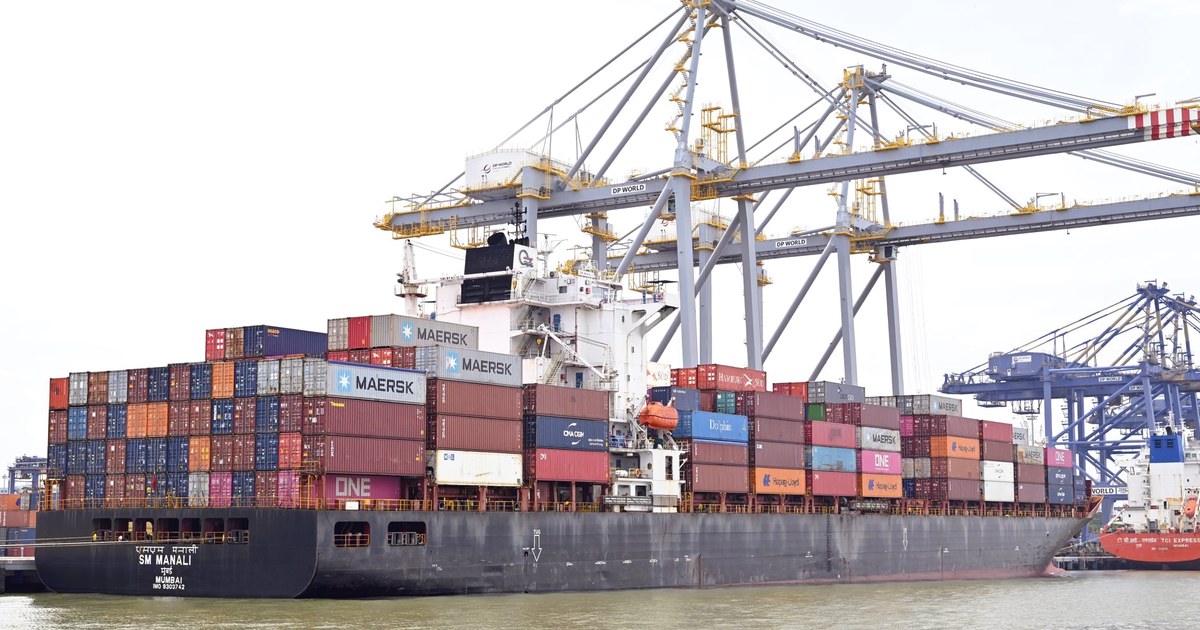 A container ship registered in Mumbai, stacked with shipping containers from Maersk and other lines, docked under gantry cranes at an Indian port