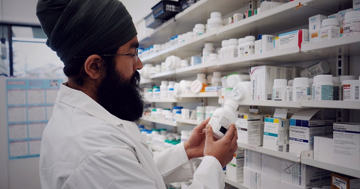 A pharmacist in a white coat and turban examines a medicine bottle against shelves stocked with packaged drugs