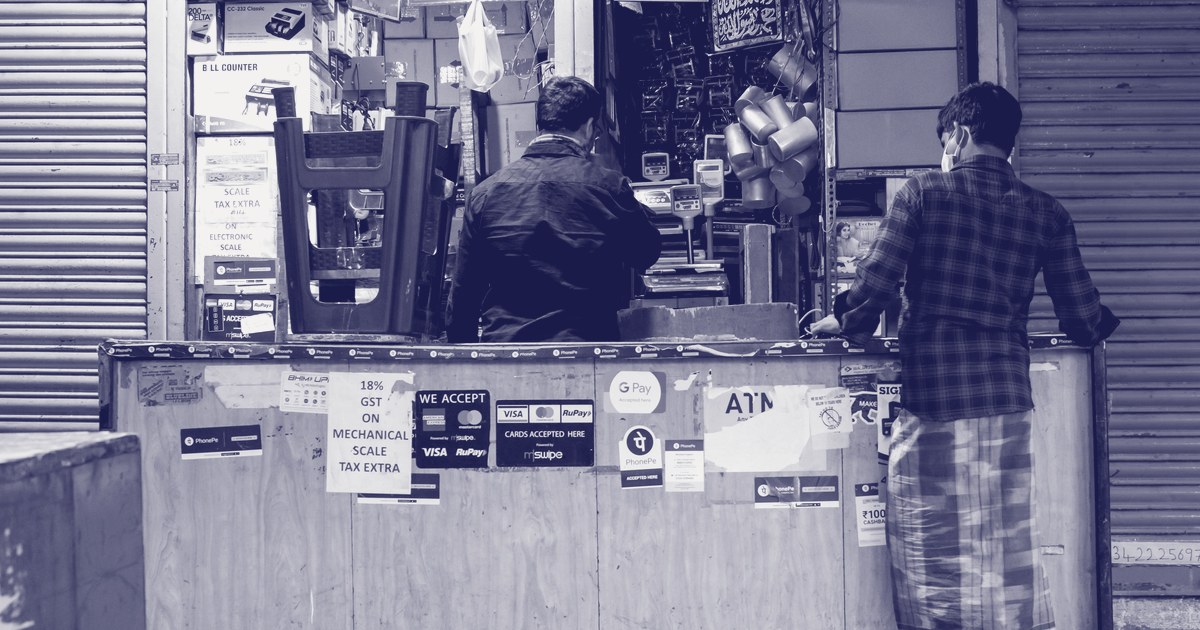 A small Indian shop counter plastered with digital-payment stickers, including GPay, PhonePe, and card acceptance signage, as two men transact at the counter