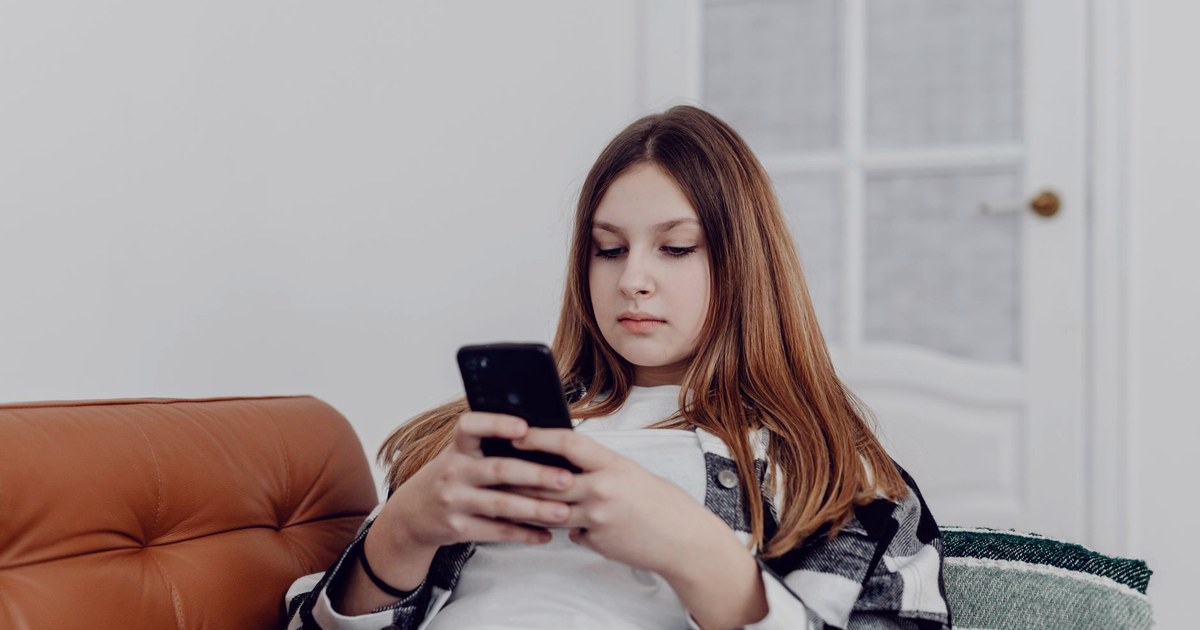 A teenage girl sitting on a sofa at home, looking at a smartphone she holds in both hands