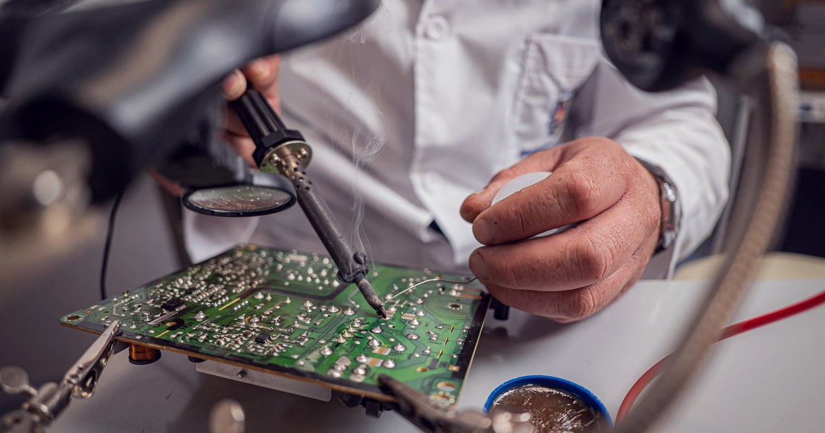 A technician in a lab coat using a soldering iron on a green printed circuit board under a lamp, with a magnifying glass and tools nearby