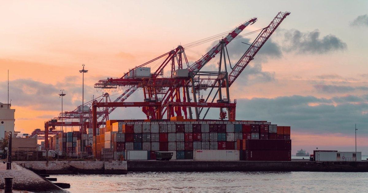 Shipping containers stacked at a port under gantry cranes at sunset