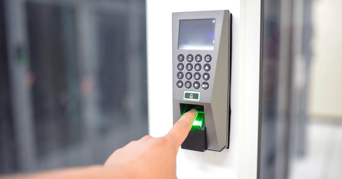 A finger pressing the green-lit fingerprint scanner on a wall-mounted biometric access panel