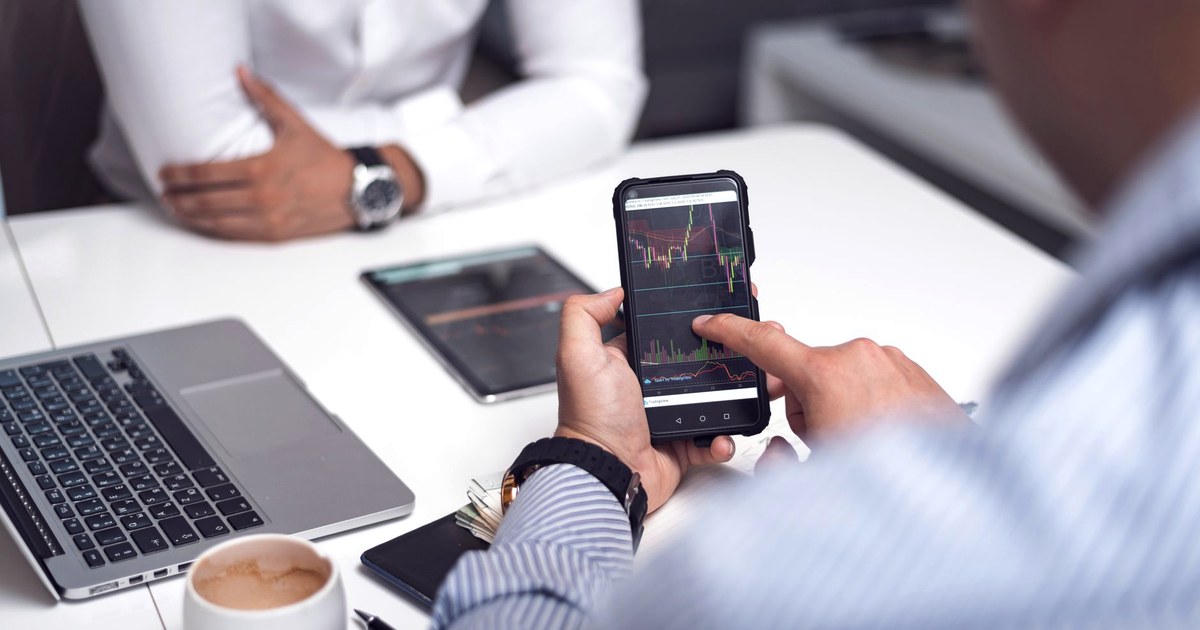 A trader reviews a cryptocurrency price chart on a smartphone during an office meeting, with a laptop and tablet on the desk