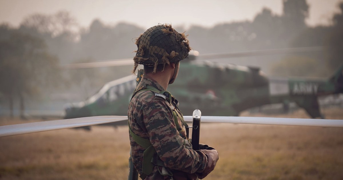 An Indian Army soldier in camouflage and a helmet stands beside a fenced perimeter, watching an olive-green Army helicopter on a misty airfield in Kolkata