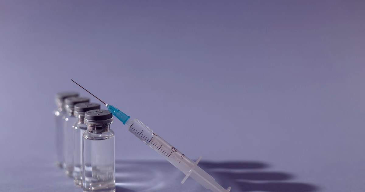 A row of glass vaccine vials with a syringe drawn from one of them, against a plain lavender-grey background