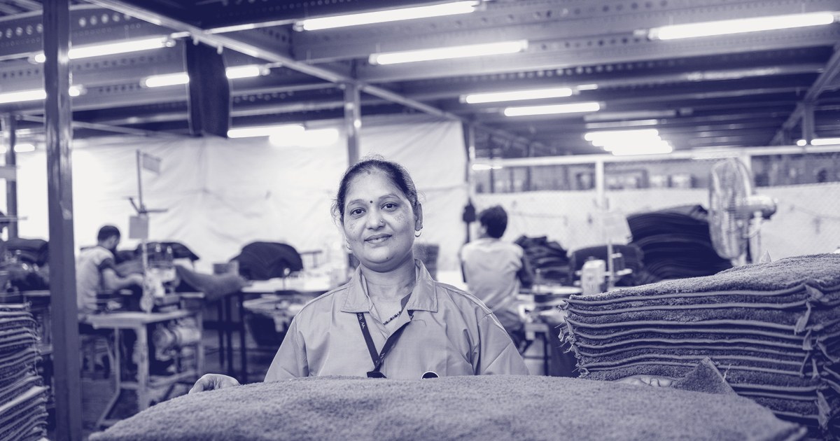 An Indian woman in a blue uniform smiling at a textile factory table, holding folded fabric, with sewing stations and fans in the background