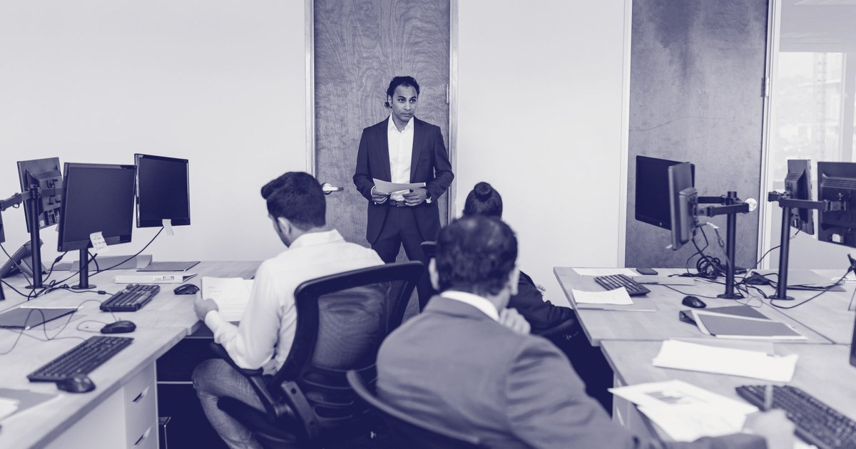 A young professional in a navy suit stands addressing three seated colleagues, two men and one woman, in an open-plan office with rows of computer monitors, shown in a navy duotone treatment