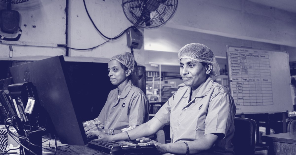 Two Indian factory workers in blue uniforms and hairnets working at computer terminals, tracking production records on a factory floor