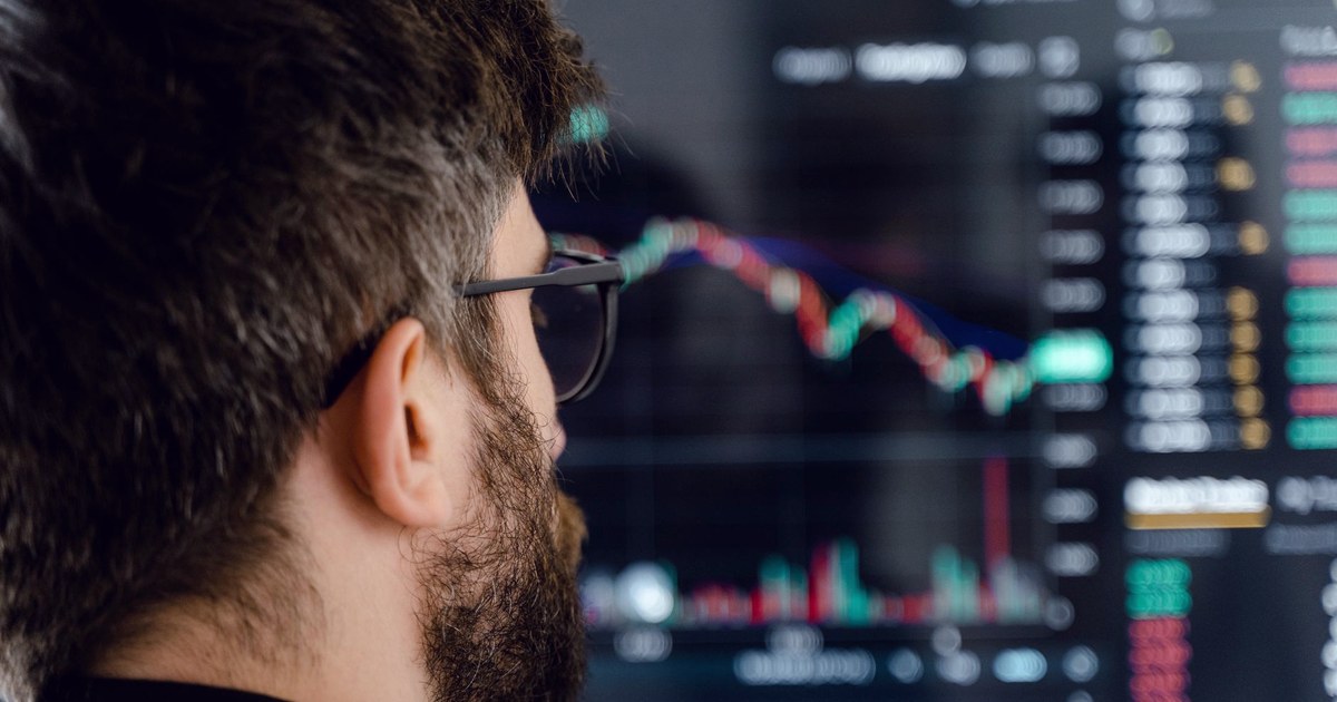 A bearded man wearing glasses examines volatile stock market charts reflected in his eyeglasses in front of a trading screen