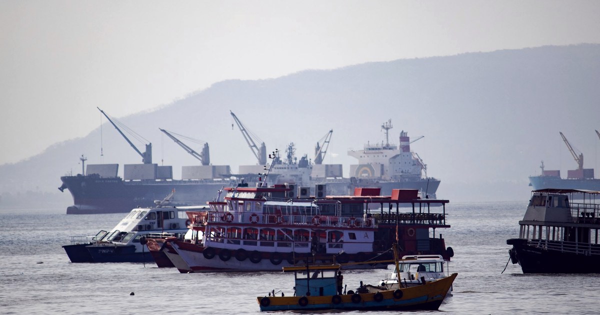 Cargo ships and harbor cranes seen across the water in Mumbai, with smaller boats in the foreground.