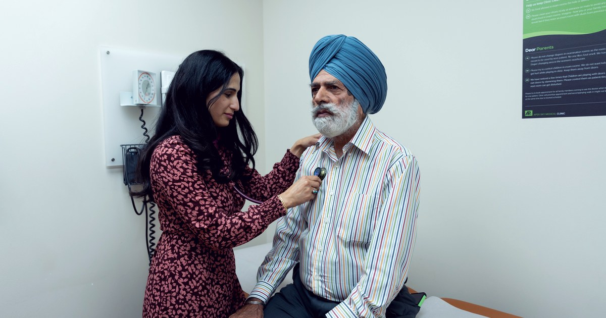 A female doctor examines a seated senior male patient with a stethoscope in a clinic exam room