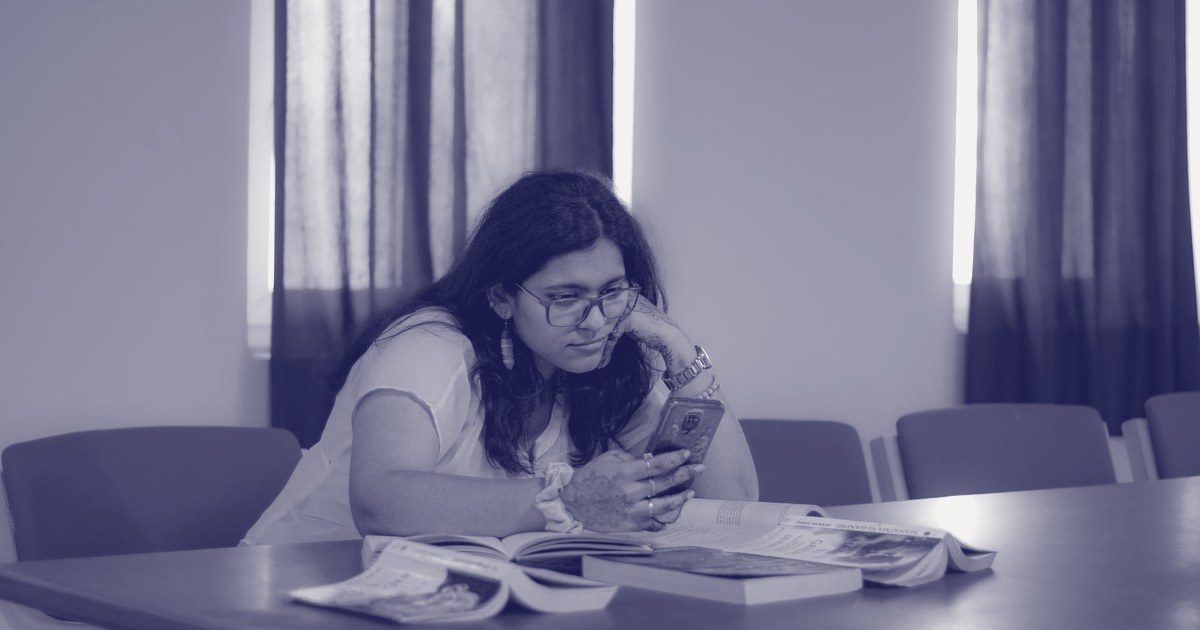 A young Indian woman with henna-decorated hands sits at a table with open books, looking at her smartphone in a classroom or library setting