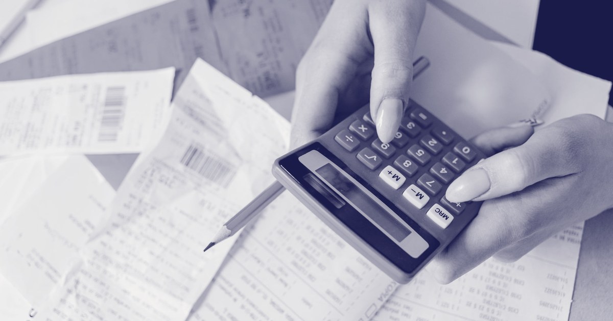 A person's hands holding a pink calculator over a spread of paper receipts and invoices on a desk