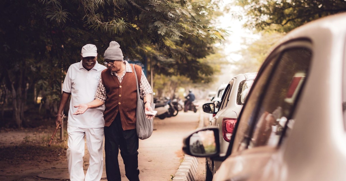 Two elderly Indian men in warm clothing walking arm in arm and laughing together on a tree-lined street, one using a walking cane