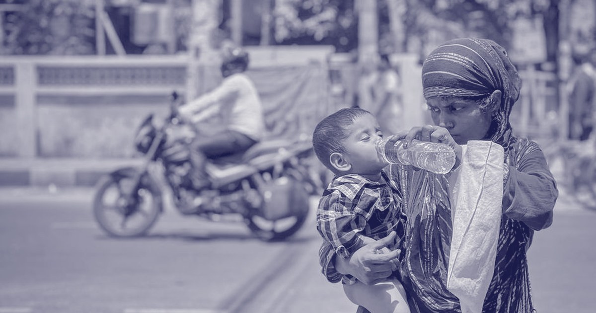 A mother shields her toddler from the heat while giving him water from a bottle on a busy Indian street, midday traffic passing behind them