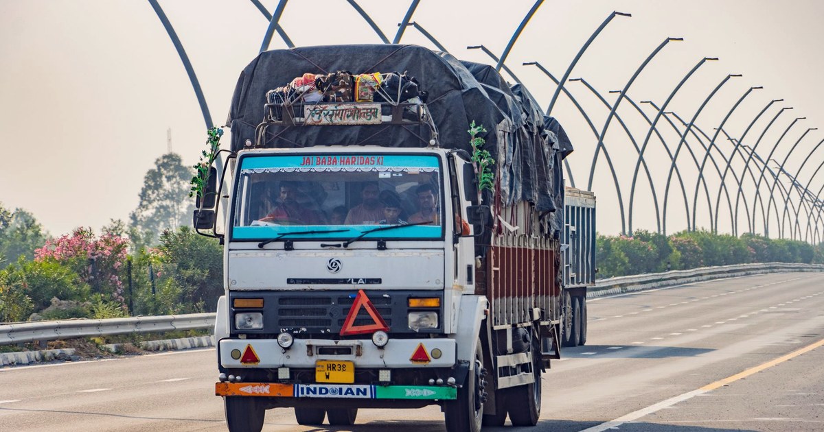 A heavily loaded truck decorated with religious motifs and carrying passengers travels down an Indian expressway, with a row of arched streetlights overhead