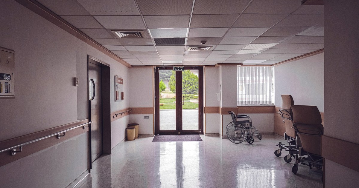 A quiet hospital corridor with a wheelchair and reclining chairs near glass doors that open onto a green garden