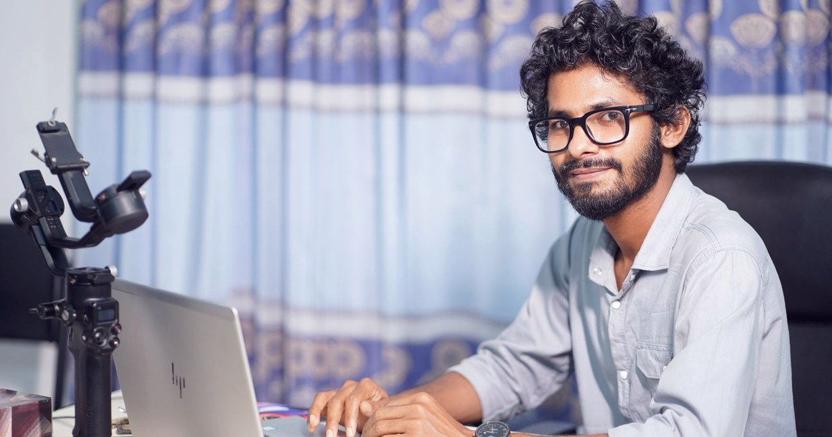 An Indian software developer wearing glasses sits at a desk with a laptop and camera rig, smiling at the camera in front of a patterned curtain