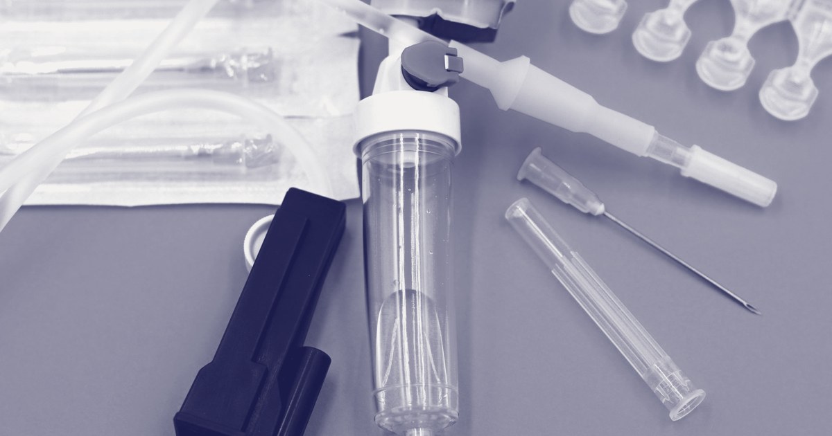 A flat-lay of unpackaged medical disposables on a blue background: IV tubing, a three-way stop cock valve, cannulas and syringe needles