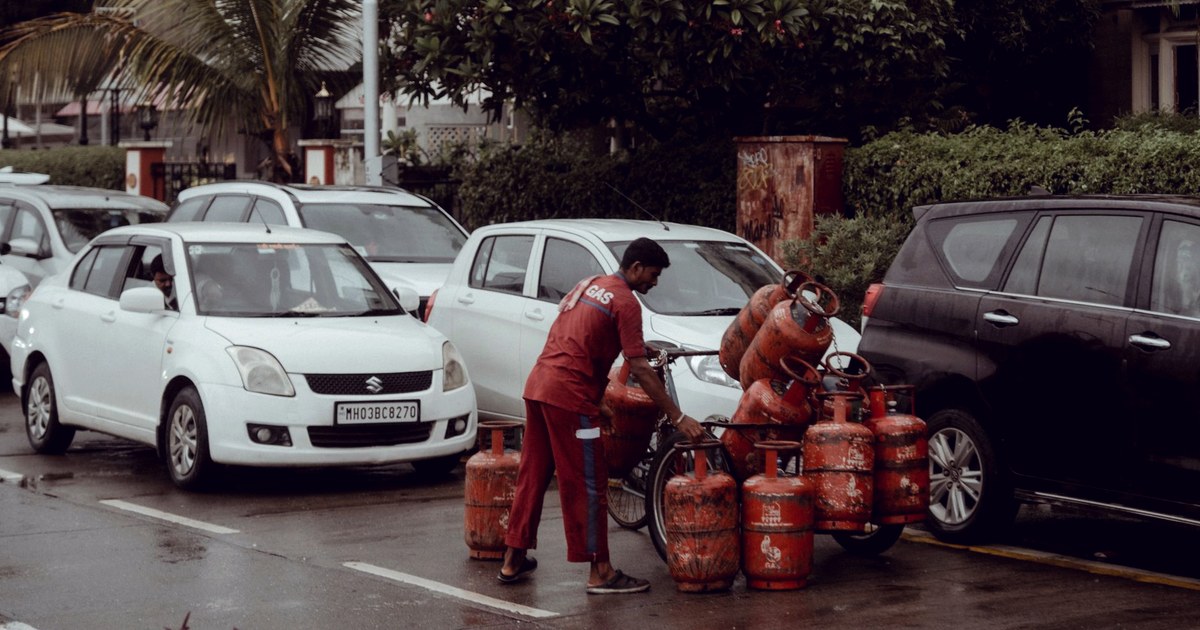 A gas delivery worker in Mumbai loads red LPG cylinders onto a bicycle cart between parked cars on a rain-slicked street