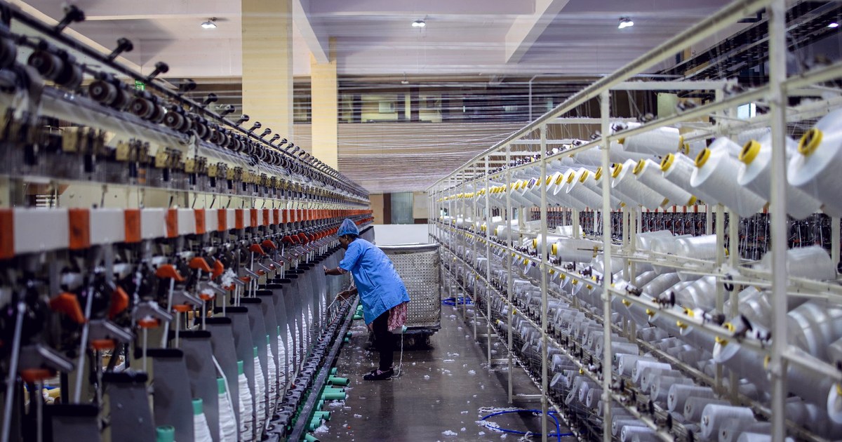 A factory worker in a blue uniform tends rows of spinning machines loaded with yarn spools on an Indian textile mill floor
